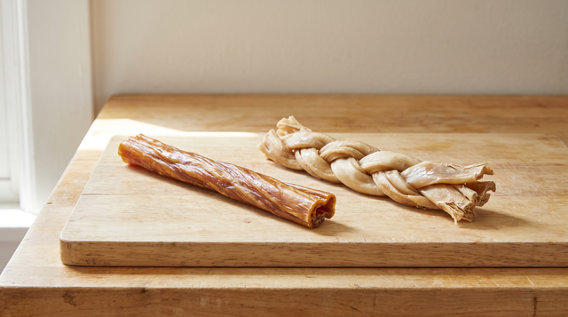 A single straight dried beef chew stick and a three-strand braided dried chew lying side by side on a wooden board on a kitchen counter